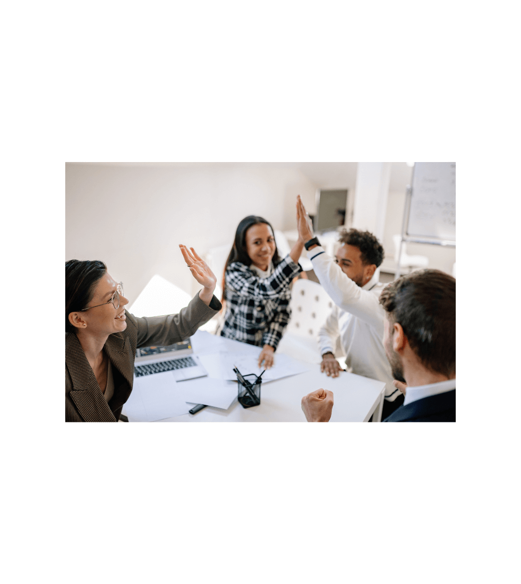 Group of diverse colleagues in an office smiling and giving high-fives around a meeting table, celebrating success.