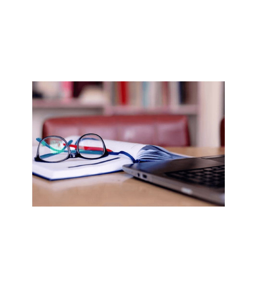 Glasses resting on an open notebook beside a laptop on a desk in a study or library setting.