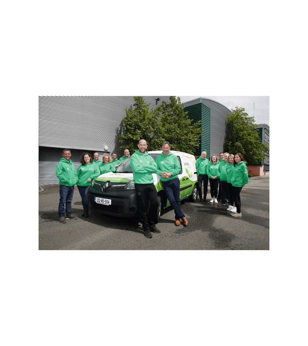 Folens team in green hoodies standing proudly around an electric delivery van outside company headquarters, promoting sustainable transport.