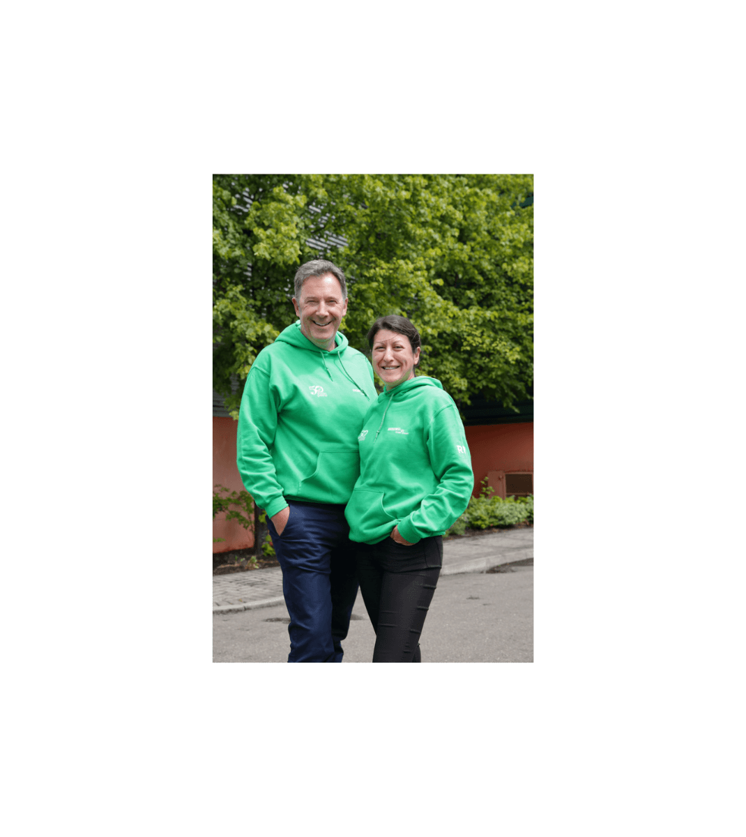 Two smiling Folens team members in matching green hoodies standing outdoors in front of trees and a red building.