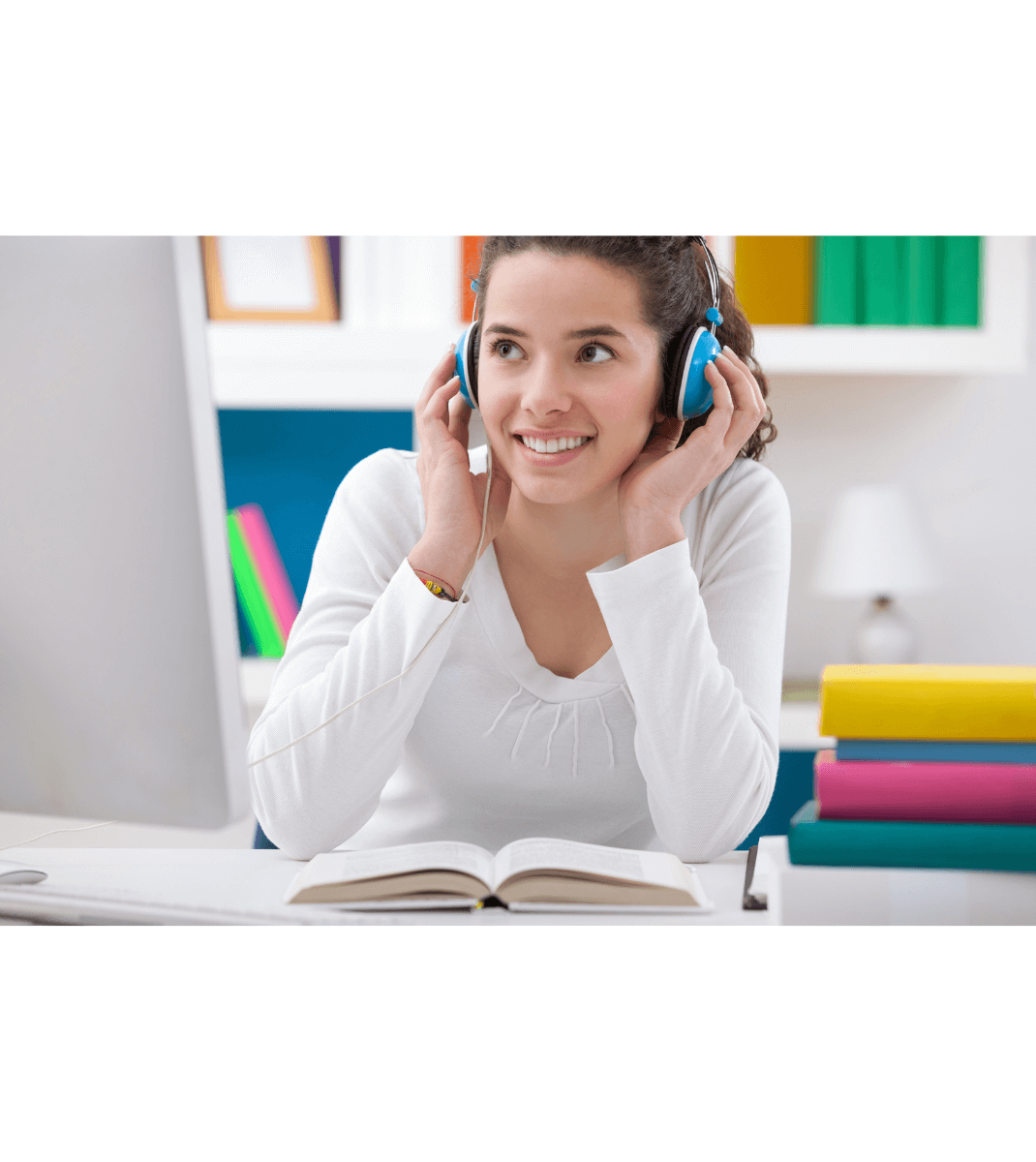 Smiling student wearing blue headphones listening to audio while studying with an open book in a colourful home workspace.