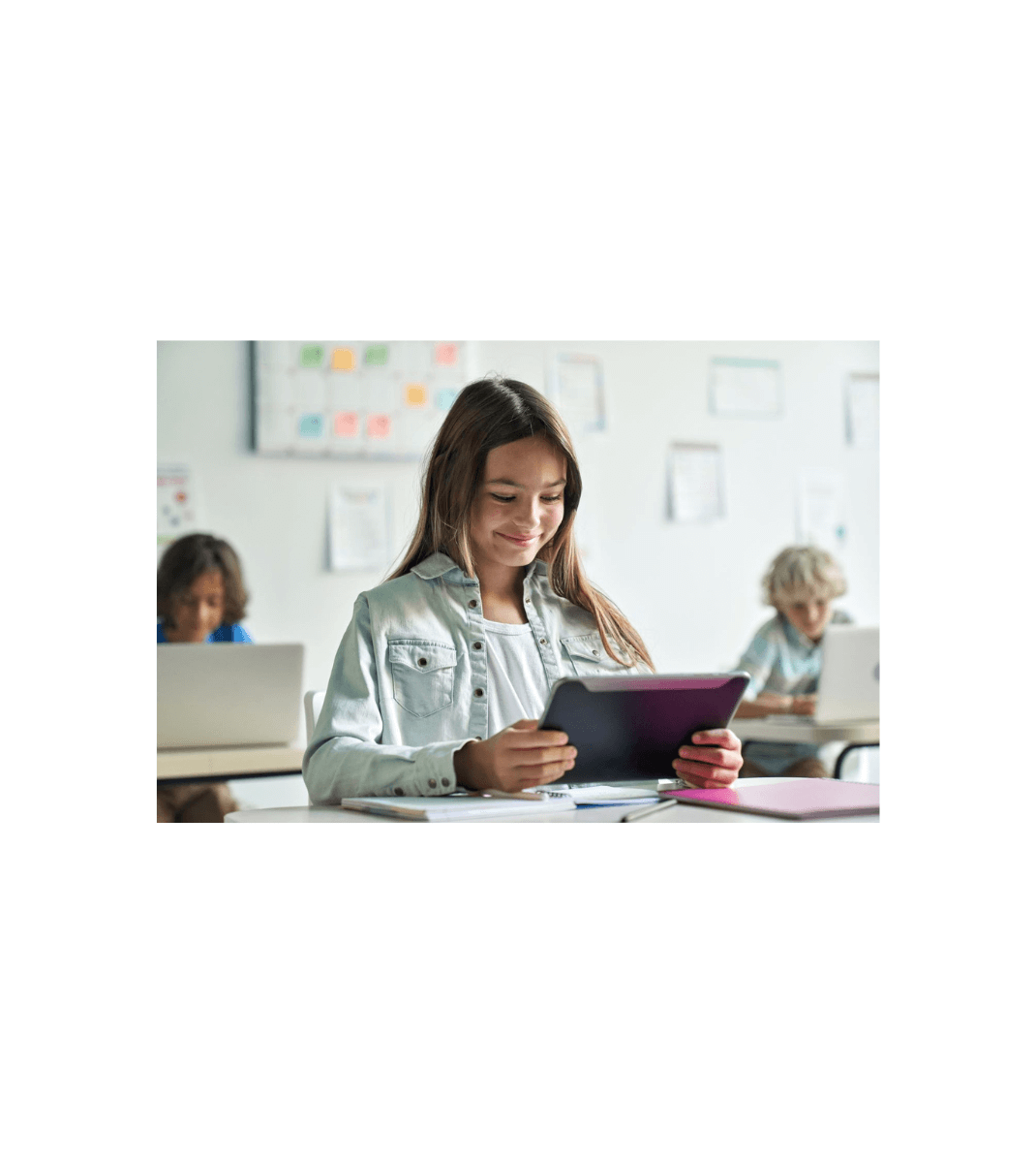 Smiling student using a tablet in a bright classroom, with other pupils working on laptops in the background.