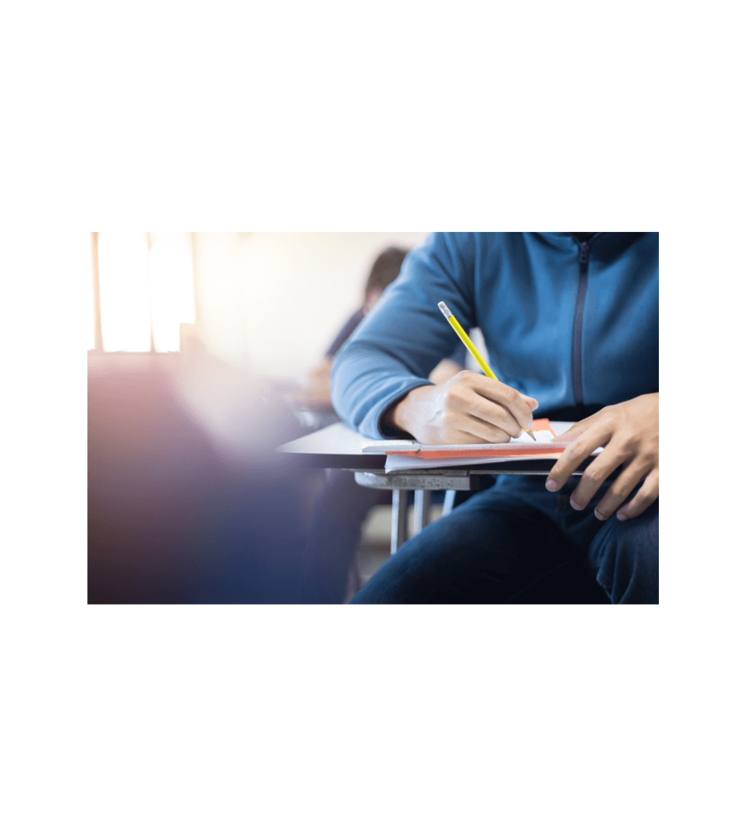 Student in blue hoodie writing with a pencil in a classroom, seated at a desk with a notebook.