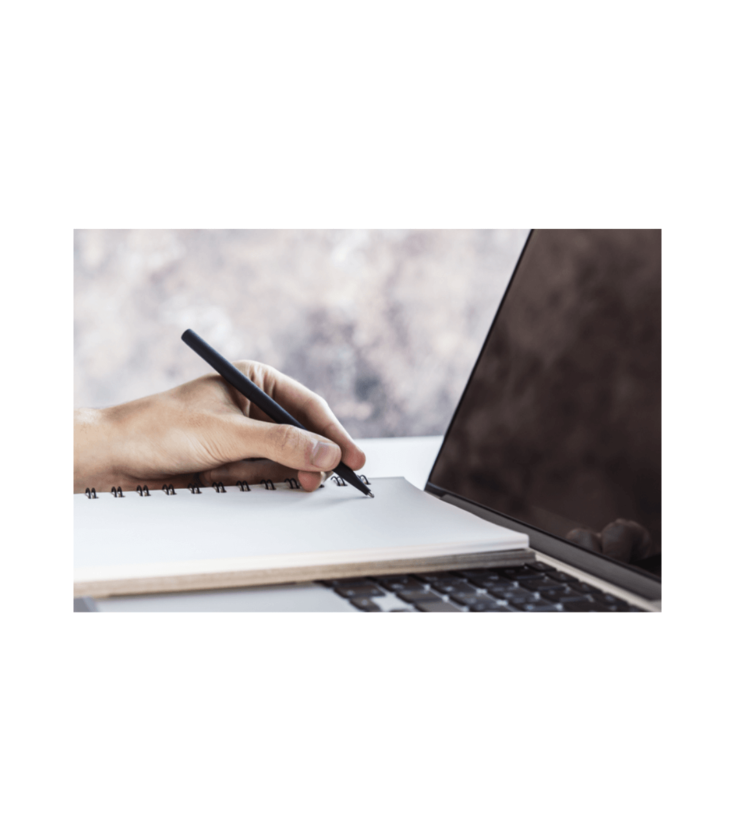 Hand writing in a spiral notebook beside an open laptop, suggesting study, research, or online learning.