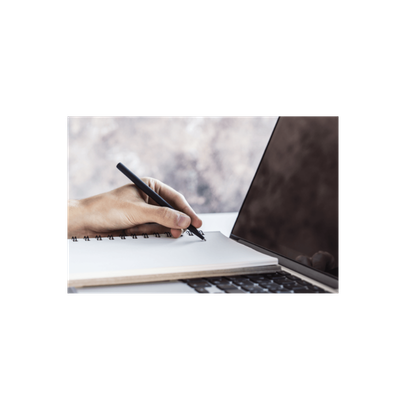 A close-up of a hand holding a black pen and writing in a white spiral notebook, with part of an open laptop visible on a light surface against a soft gray background.